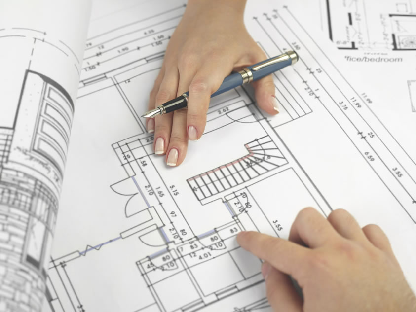 Two hands on architectural blueprints, one marking with a pencil, emphasizing details and measurements on a building plan.