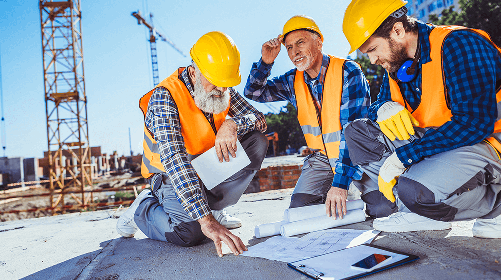 Three construction workers in hard hats and high visibility vests discuss plans on a blueprint at a construction site with cranes in the background.