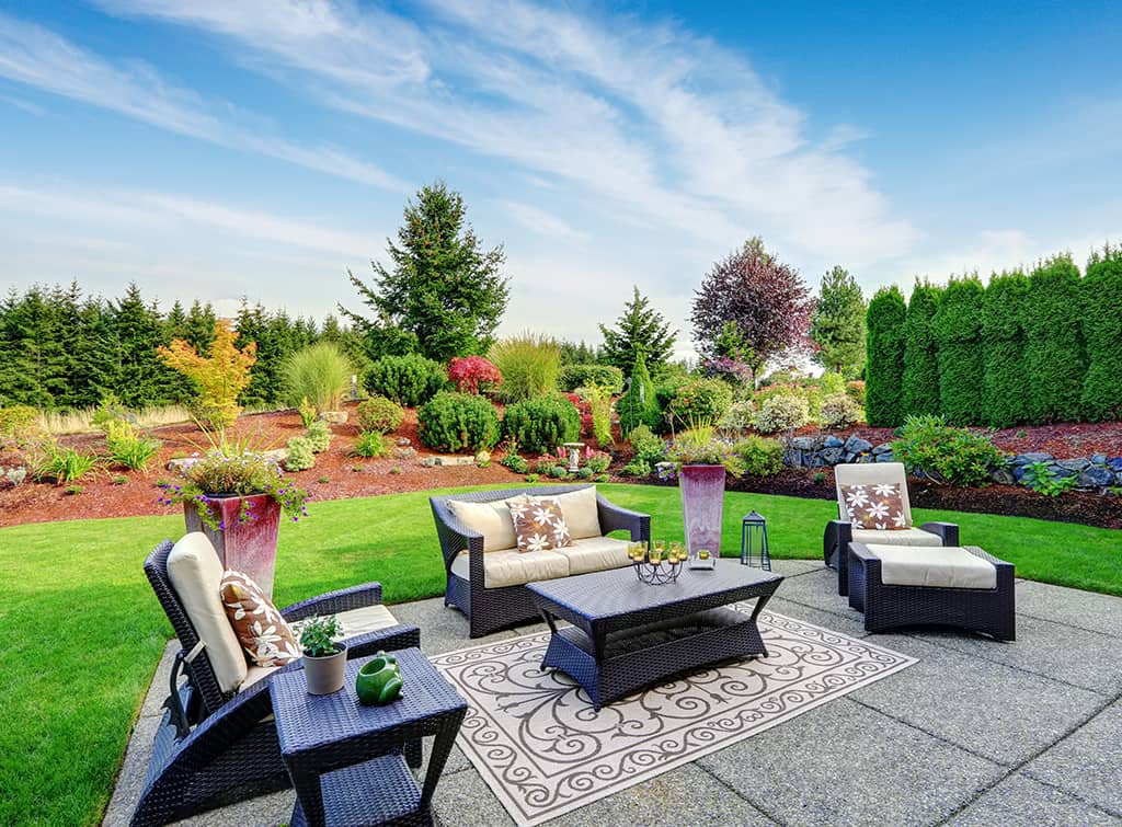 A landscaped garden with lush green lawn, assorted plants, and trees; furnished with a rattan patio set including sofas and a coffee table, under a clear blue sky.