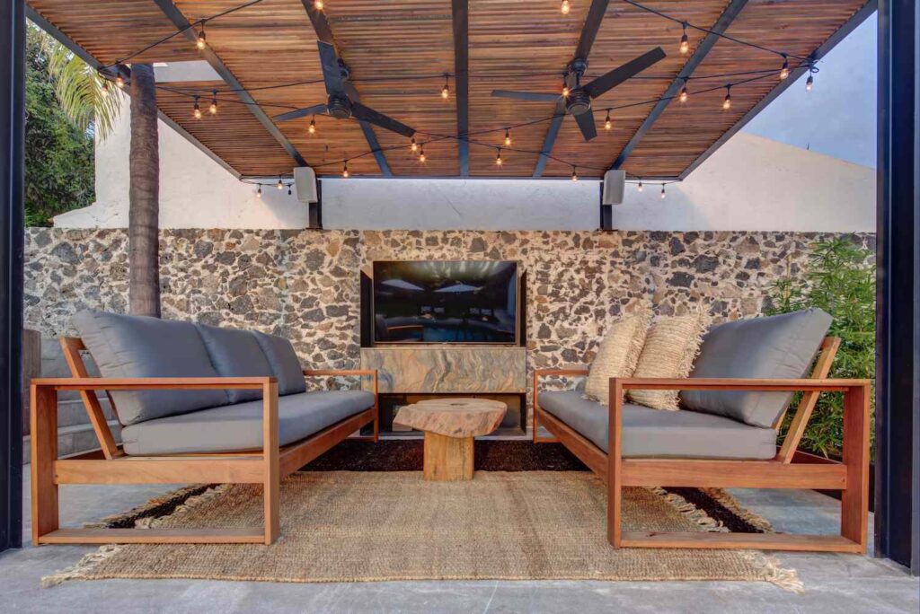 Outdoor patio with two wooden armchairs with gray cushions, a small wooden coffee table, and a stone wall with a mounted television. area is covered and includes ceiling fans.